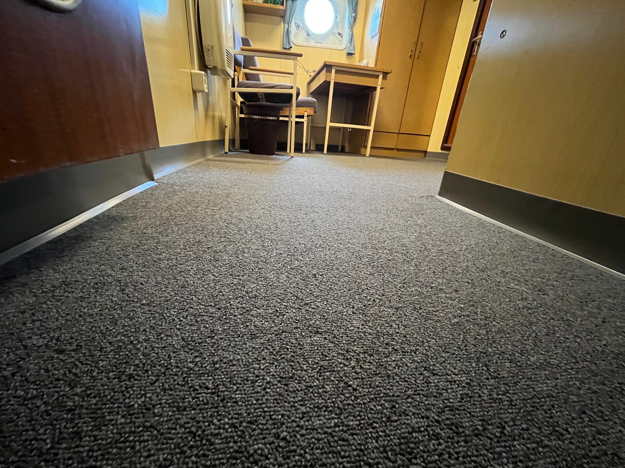 Szara wykładzina dywanowa w pomieszczeniu z biurkiem, krzesłami i oknem okrągłym w tle, widok z niskiej perspektywy.