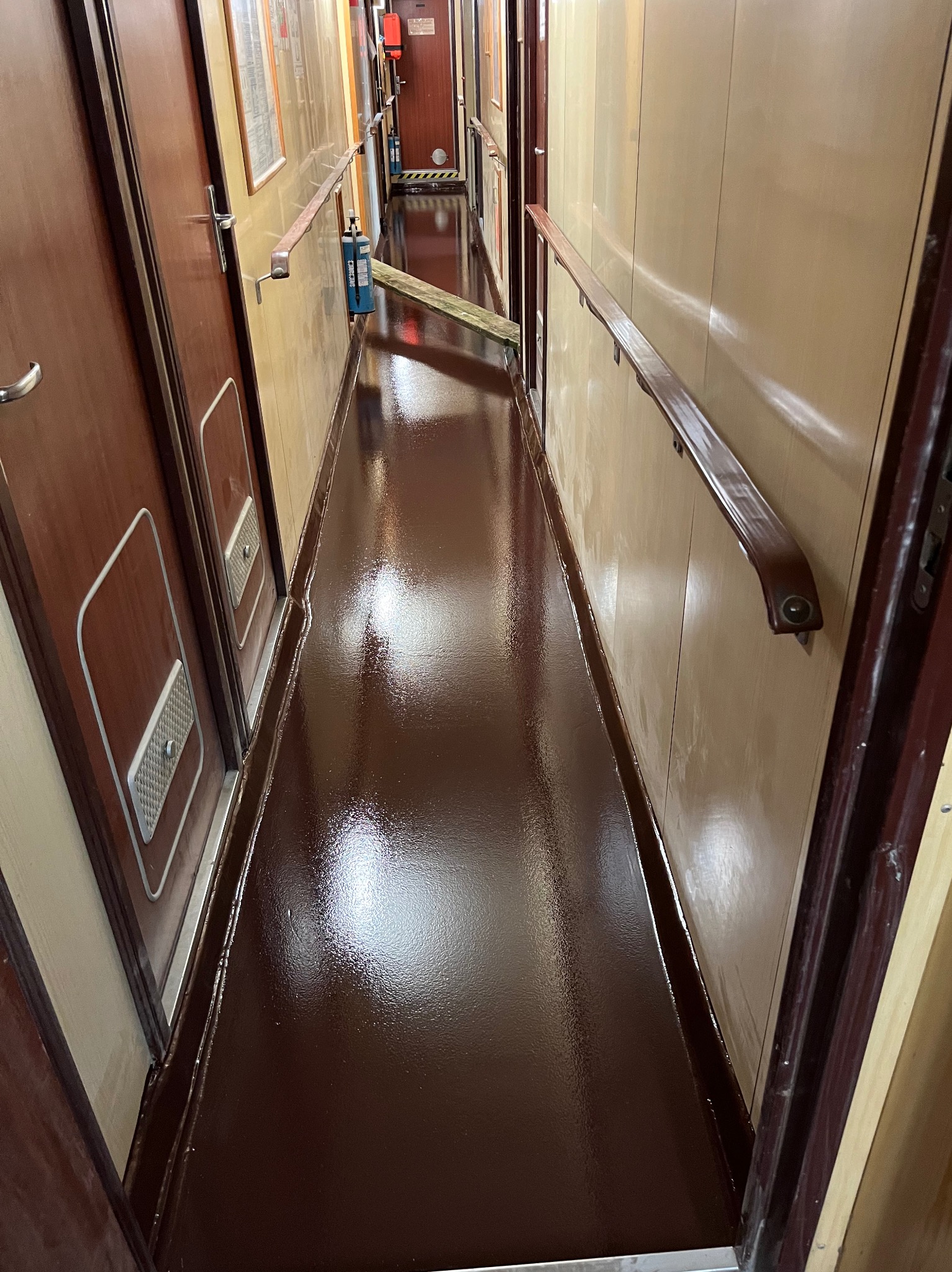Błyszcząca, brązowa podłoga w wąskim korytarzu z drewnianymi drzwiami i poręczą, w tle widoczne narzędzia i butla z gazem.