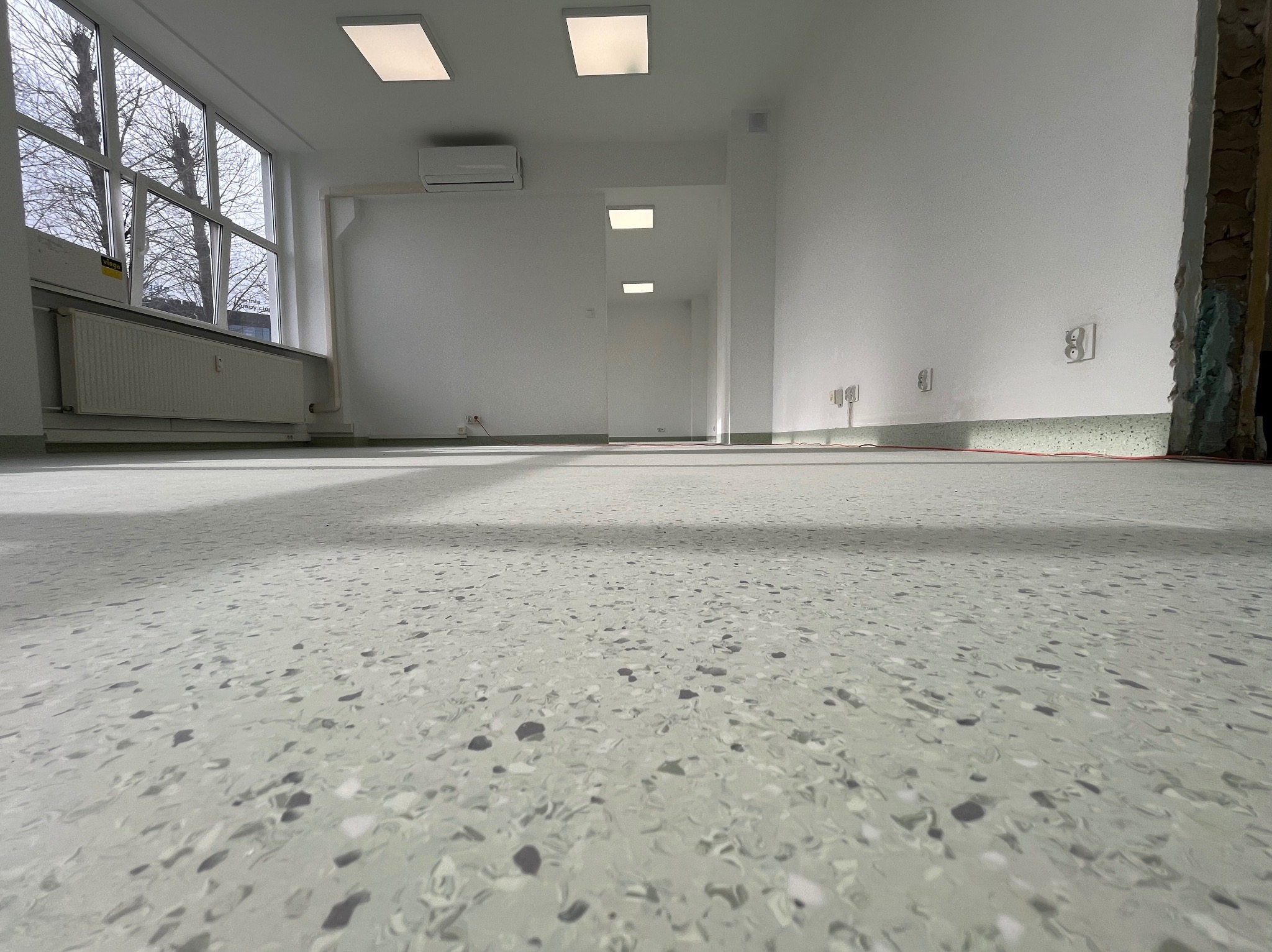 Jasnoszara, wzorzysta podłoga w pustym, jasnym pomieszczeniu z oknem i grzejnikiem, widoczne gniazdka elektryczne i fragment ściany w trakcie wykończenia.