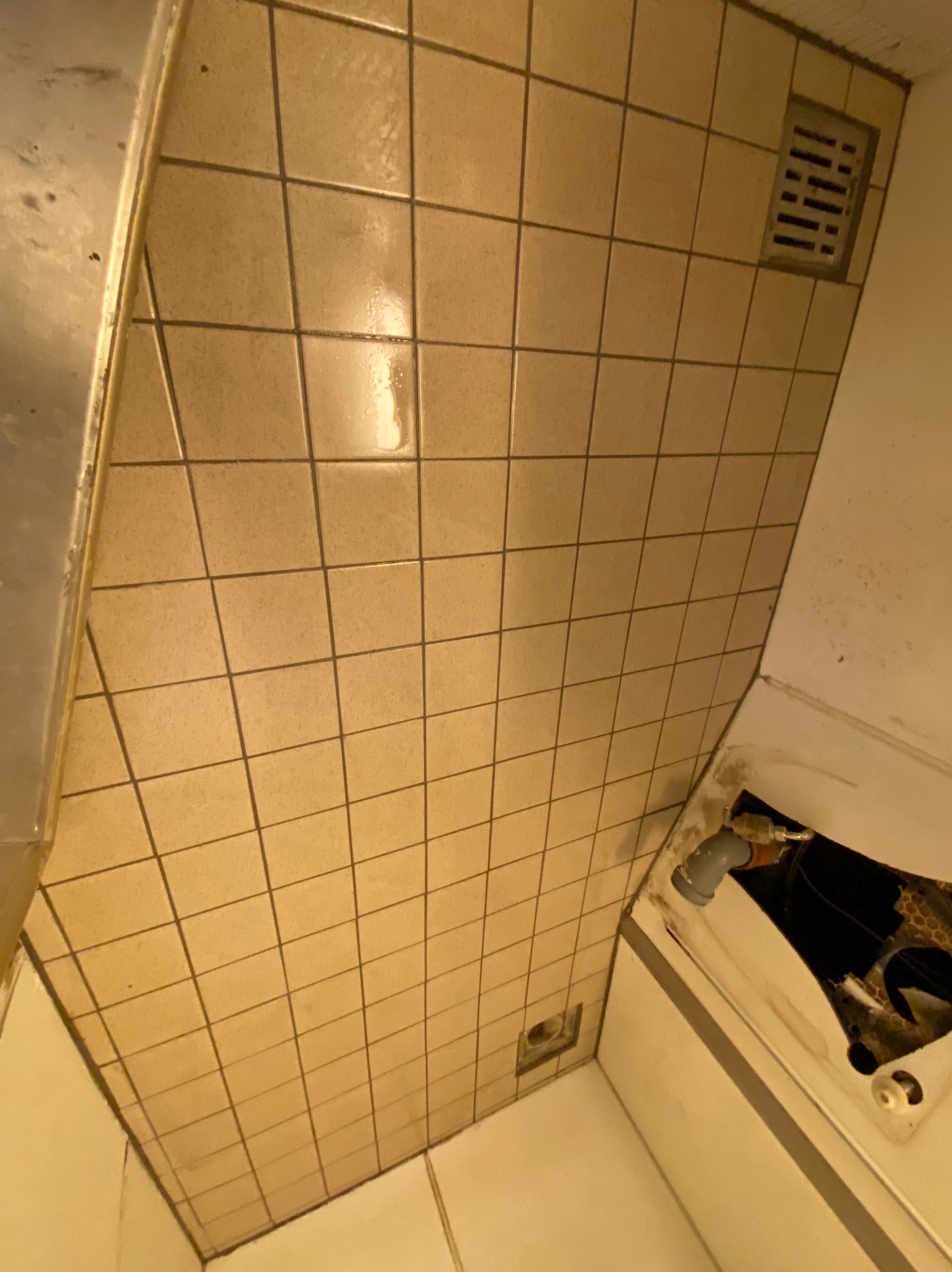 Żółte, kwadratowe płytki ceramiczne na ścianie w łazience, widoczna kratka wentylacyjna, fragment wanny z uszkodzeniami i instalacją hydrauliczną.