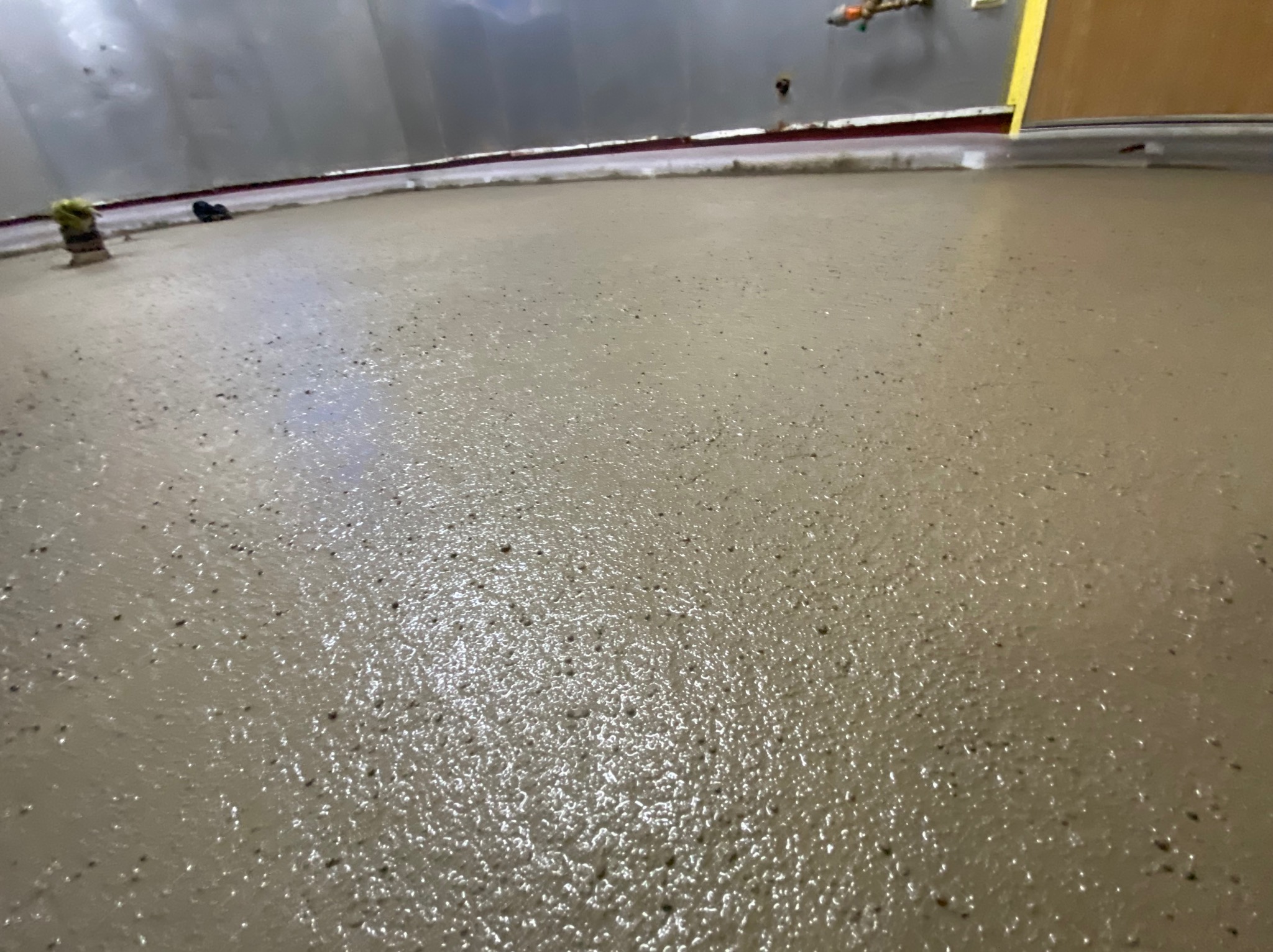 Świeżo wylana, wilgotna posadzka żywiczna w szarym odcieniu, widoczne detale struktury powierzchni i odblaski światła, przy ścianie listwa przypodłogowa.