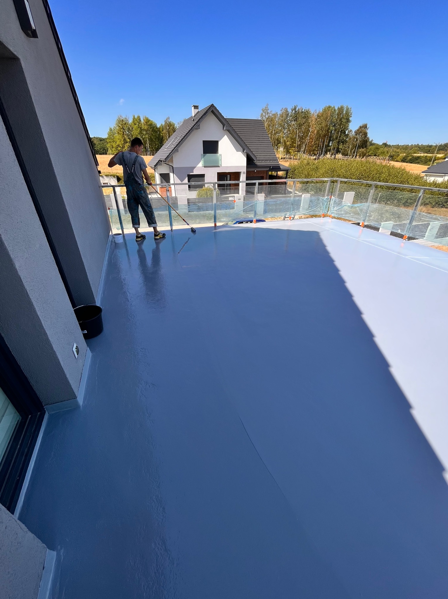 Aplikacja szarej, błyszczącej powłoki na balkonie z szklanymi balustradami; w tle dom jednorodzinny i błękitne niebo.