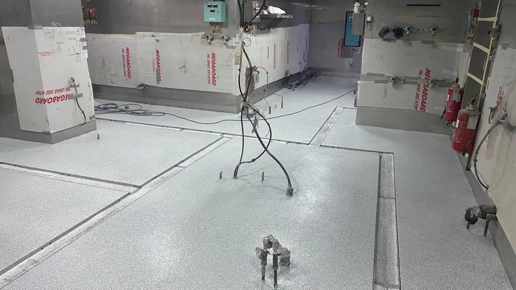 Szara posadzka w hali przemysłowej z widocznymi dylatacjami i sprzętem montażowym, ściany wyłożone płytami ochronnymi.