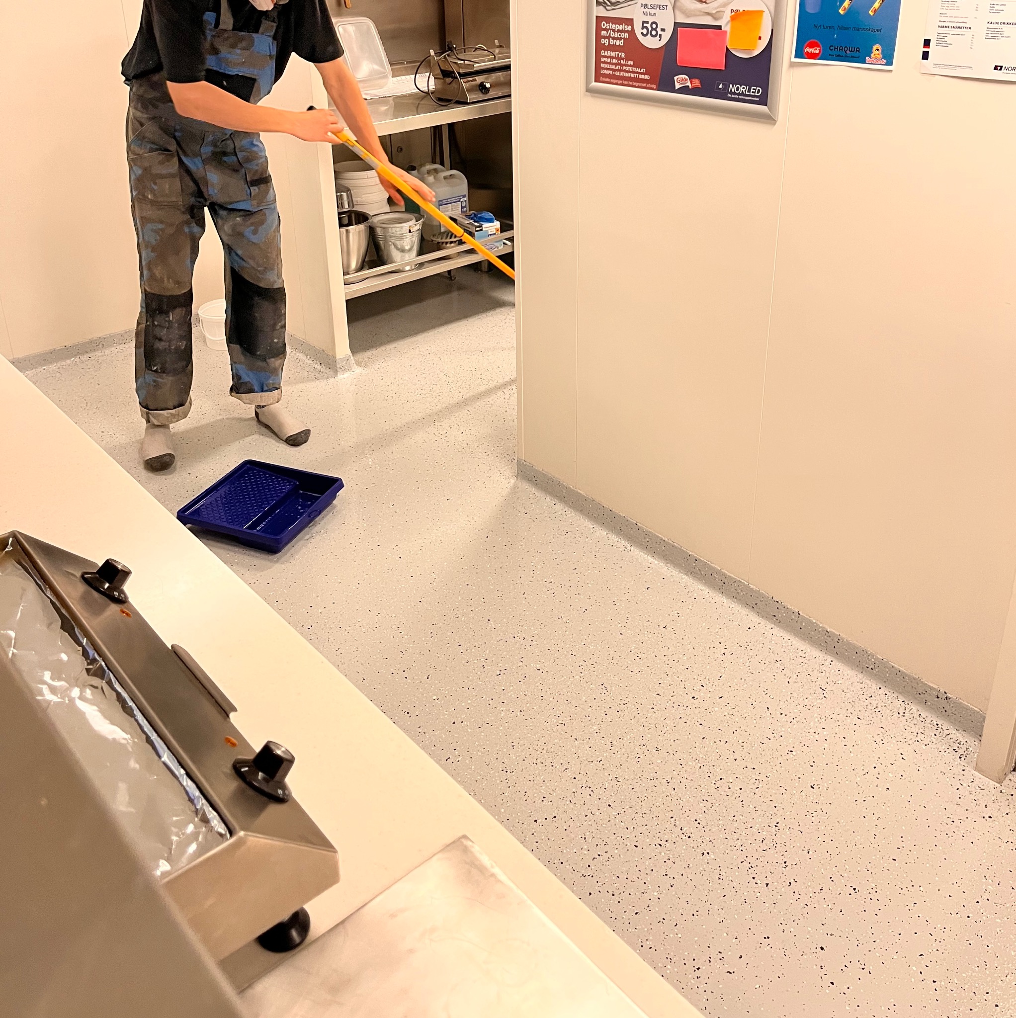 Pracownik w kombinezonie malarskim rozprowadza wałkiem jasną posadzkę z ciemnymi drobnymi plamkami w pomieszczeniu z metalowymi regałami i niebieską kuwetą malarską na podłodze.