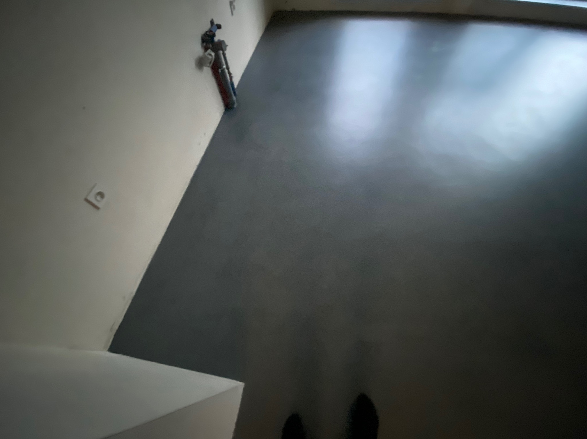 Szara, błyszcząca podłoga w nowym pomieszczeniu, widoczna instalacja hydrauliczna przy ścianie, fragment buta w rogu kadru.
