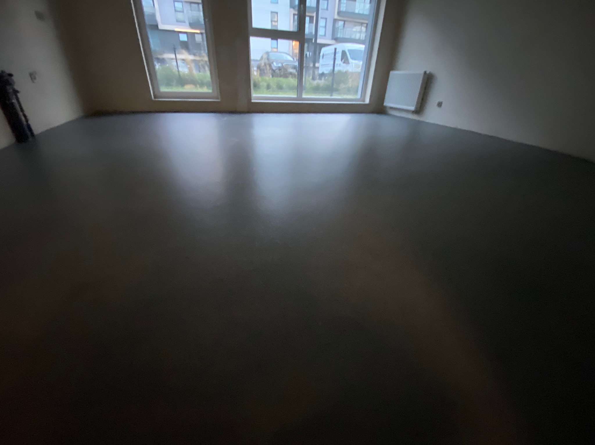 Wnętrze pokoju z nową, ciemną, błyszczącą podłogą, widok na okna z budynkami na zewnątrz.