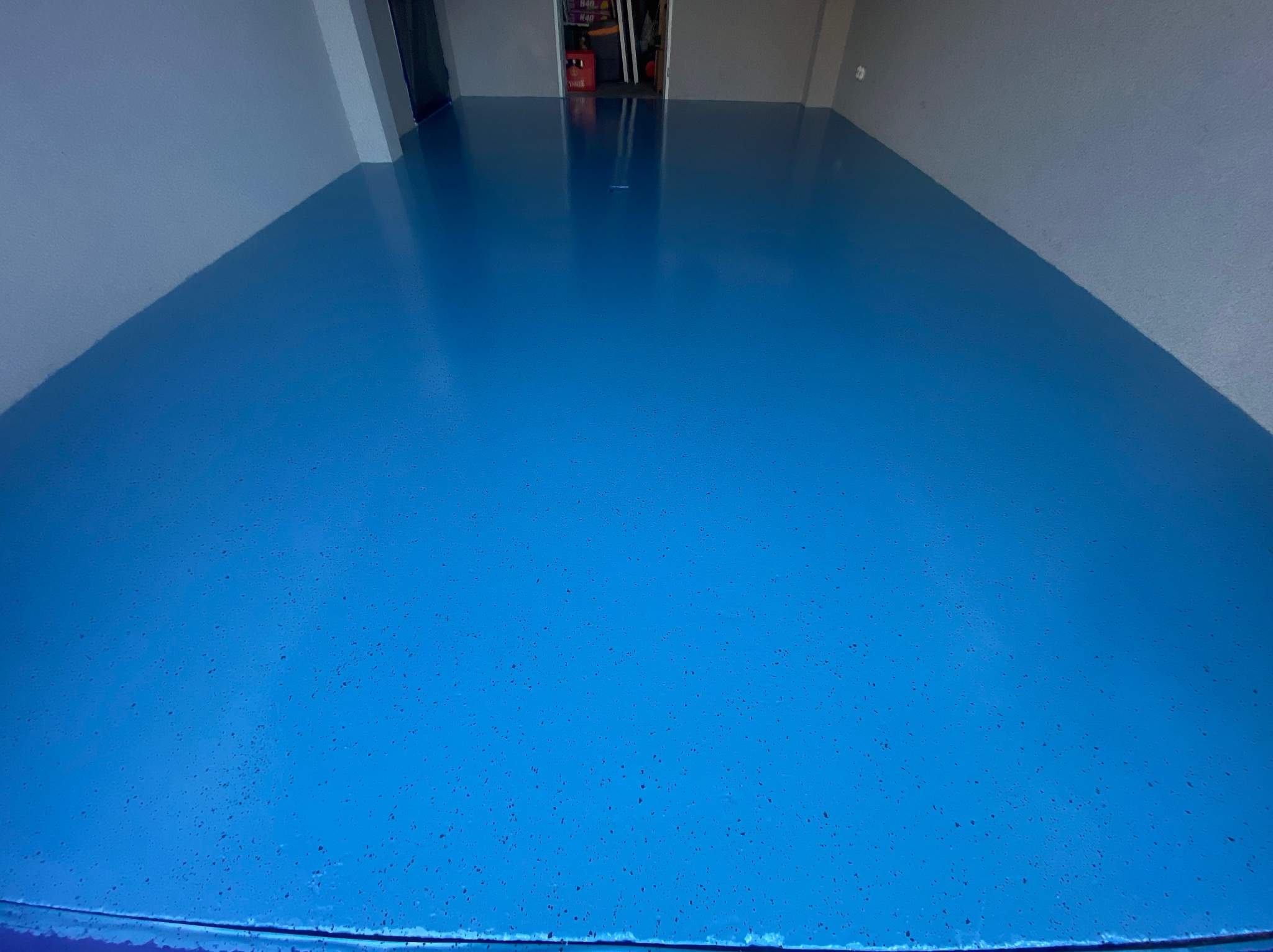 Błękitna posadzka żywiczna z delikatnym, ciemniejszym wzorem w garażu, ściany w odcieniach szarości, widoczny fragment drzwi w tle.
