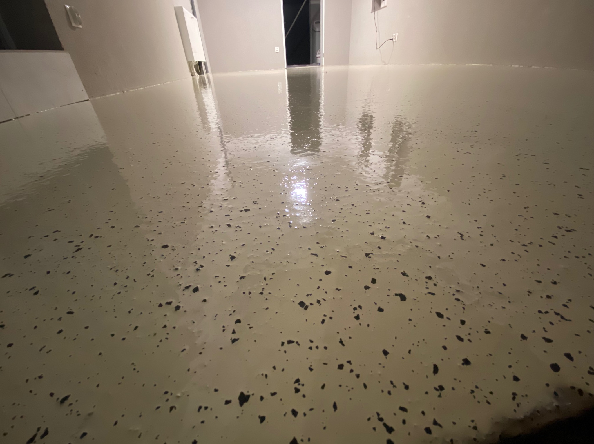 Jasna, błyszcząca podłoga w pomieszczeniu z czarnymi, nieregularnymi wtrąceniami, widoczne ślady po wałku, w tle białe ściany i otwarte drzwi.