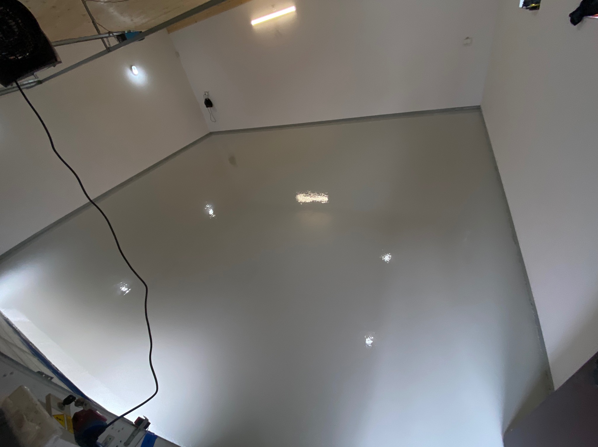 Świeżo wylana, jasnoszara, gładka powierzchnia podłogi w pomieszczeniu, z widocznymi odbiciami światła i elementami wentylacji przy suficie.