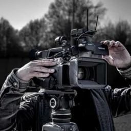 Profesjonalna kamera Sony XDCAM na statywie, z operatorem w skórzanej kurtce w trakcie wymiany karty pamięci, na rozmytym tle drzew.