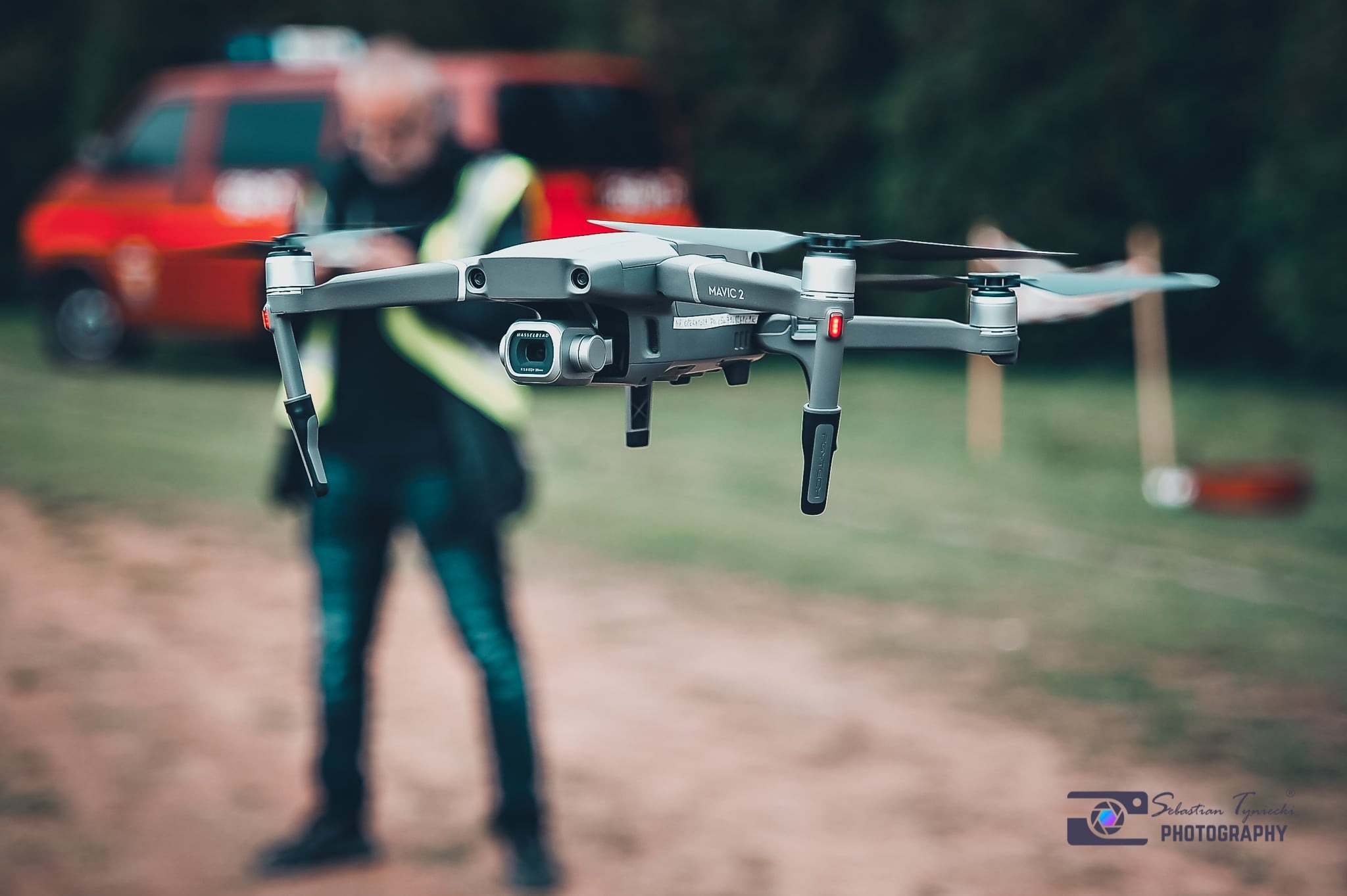 Szary dron Mavic 2 w locie, w tle rozmyty strażak i wóz strażacki, ziemista droga.