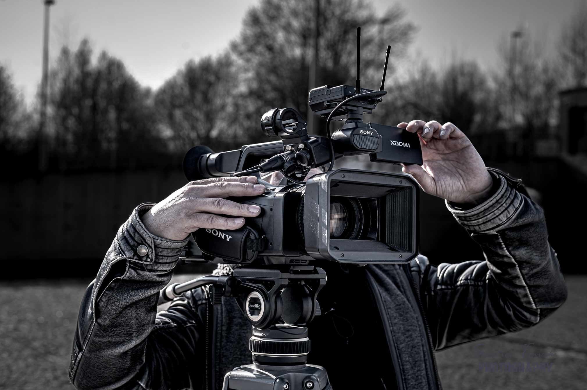 Profesjonalna kamera Sony XDCAM na statywie, z operatorem w skórzanej kurtce w trakcie wymiany karty pamięci, na rozmytym tle drzew.