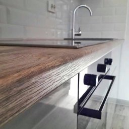 Czystosc Project - Blat kuchenny z imitacją drewna, wbudowana płyta grzewcza, chromowana bateria i białe kafelki na ścianie w tle.