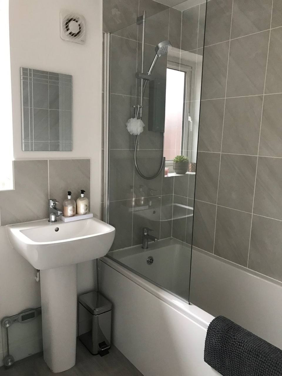 Umywalka na postumencie z baterią, obok kosz na śmieci ze stali nierdzewnej, w tle wanna z szklanym parawanem i prysznicem, szare płytki ścienne, lustro z dekoracyjnymi paskami