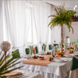 Uroczysty st&oacute;ł z białym obrusem, brązowym bieżnikiem, białymi talerzami, szklankami z serwetkami w zielone liście, dekoracjami z sukulent&oacute;w i wysoką kompozycją z zielonych roślin w złotym wazonie.