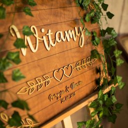 Opus Dorota Kaak - Drewniana tablica powitalna z napisem 'Witamy', imionami 'Patrycja & Marcin' i datą '21.05.2022', ozdobiona zielonymi girlandami, ustawiona na drewnianym stojaku, widoczny fragment fotela w tle.