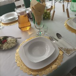 Opus Dorota Kaak - Elegancko nakryty stół z białym obrusem, złotymi podkładkami pod talerze, białymi naczyniami, dzbankiem z napojem, sałatką, winogronami i butelkami napojów.