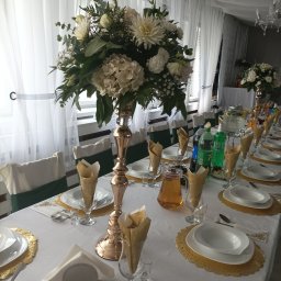 Opus Dorota Kaak - Uroczysty stół z białym obrusem, złotymi podkładkami, elegancką zastawą, kwiatowymi dekoracjami w wysokich, złotych wazonach oraz napojami. Krzesła z zielonymi akcentami. W tle białe, zwiewne firany.