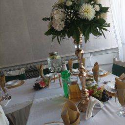 Opus Dorota Kaak - Elegancko nakryty stół z białym obrusem, złotymi podkładkami, wysokim wazonem z białymi kwiatami, szklanymi naczyniami z napojami, owocami i serwetkami w szklankach, przygotowany na przyjęcie.