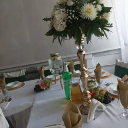 Elegancko nakryty stół z białym obrusem, złotymi podkładkami, wysokim wazonem z białymi kwiatami, szklanymi naczyniami z napojami, owocami i serwetkami w szklankach, przygotowany na przyjęcie.