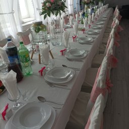Opus Dorota Kaak - Długi stół bankietowy, nakryty białym obrusem, z białymi talerzami, sztućcami, szklankami, dzbankami z napojami i dekoracjami kwiatowymi w wysokich wazonach. Krzesła udekorowane różową wstążką.