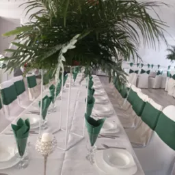 Uroczysty stół z białym obrusem, nakryciami i zielonymi serwetkami, ozdobiony wysokimi kompozycjami z tropikalnych liści i srebrnymi świecznikami, krzesła z zielonymi szarfami.