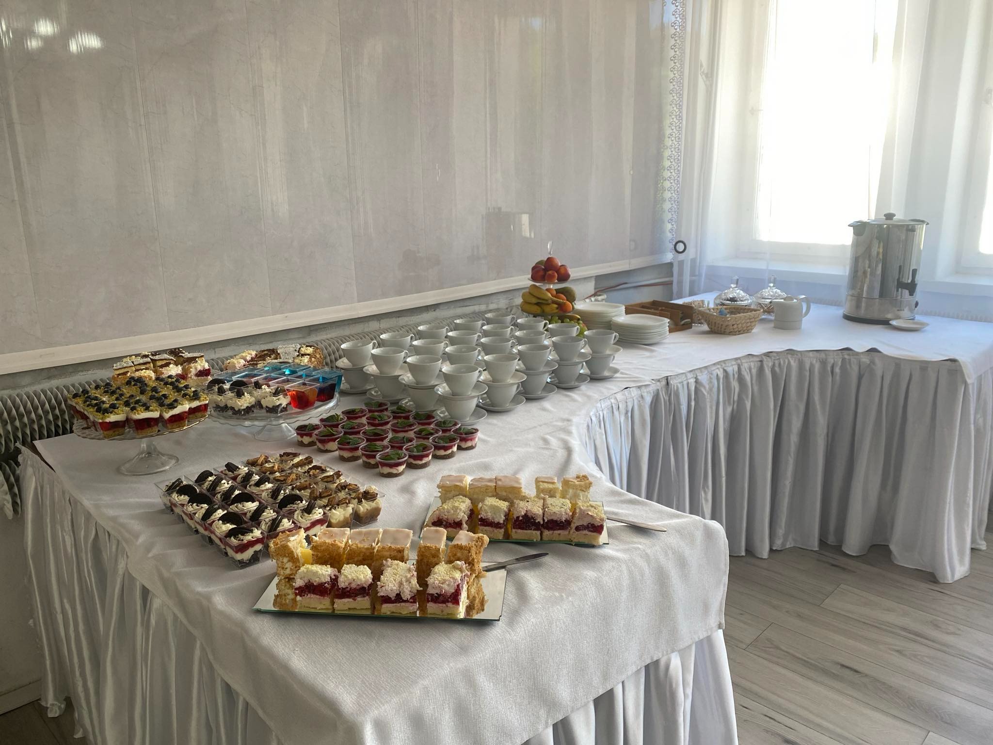Stół cateringowy z deserami: ciastka z owocami, mini deserki w szklankach, piramida z owoców, stosy białych filiżanek i podgrzewacz do wody, wszystko przykryte białym obrusem z falbanami.