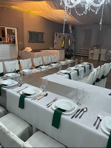 Uroczysta sala z białymi krzesłami i stołami w kształcie litery U, nakrytymi białymi obrusami z delikatnym wzorem i zielonymi serwetkami. W tle widoczna dekoracja z balonów w kształcie cyfry 10.