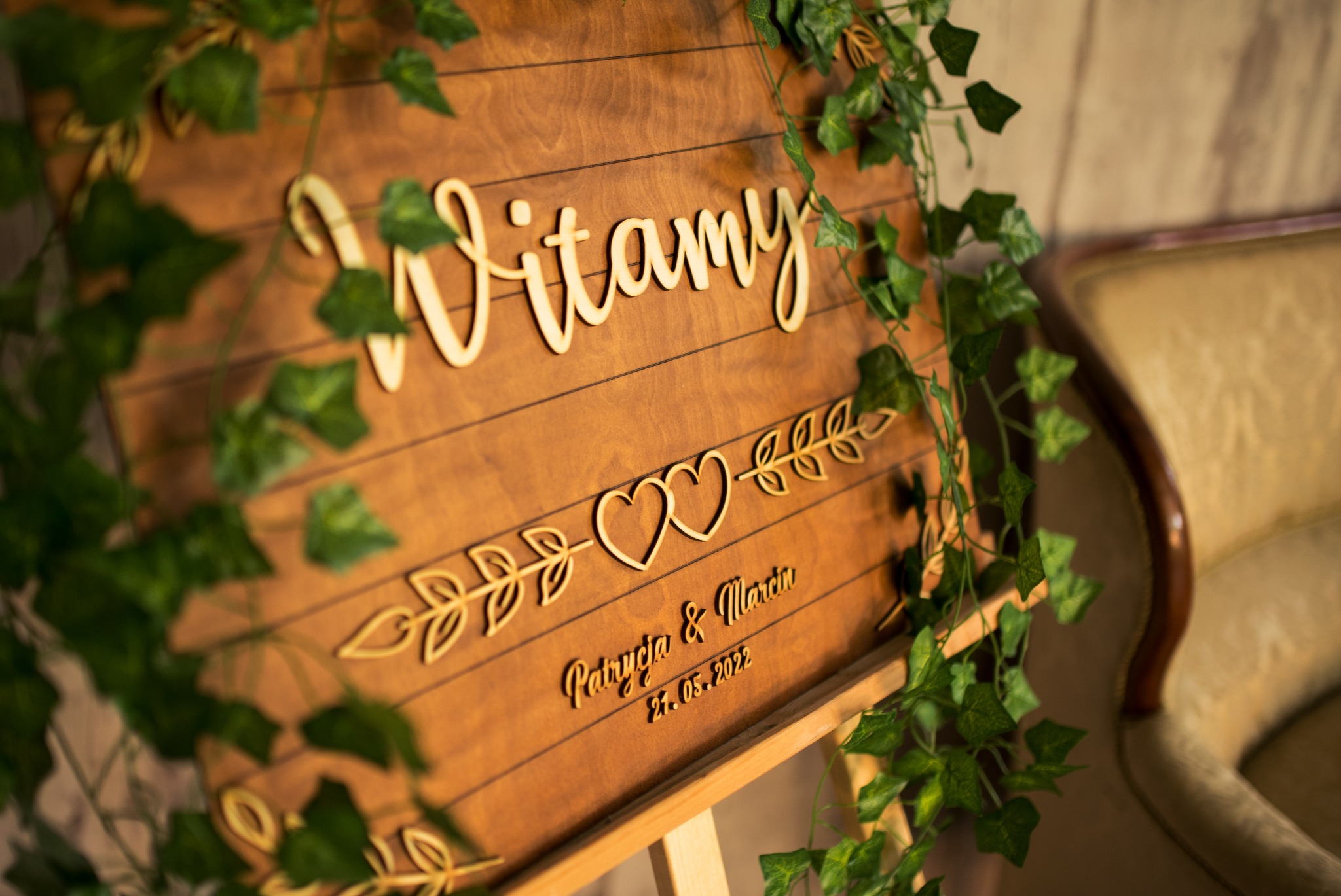 Drewniana tablica powitalna z napisem 'Witamy', imionami 'Patrycja & Marcin' i datą '21.05.2022', ozdobiona zielonymi girlandami, ustawiona na drewnianym stojaku, widoczny fragment fotela w tle.