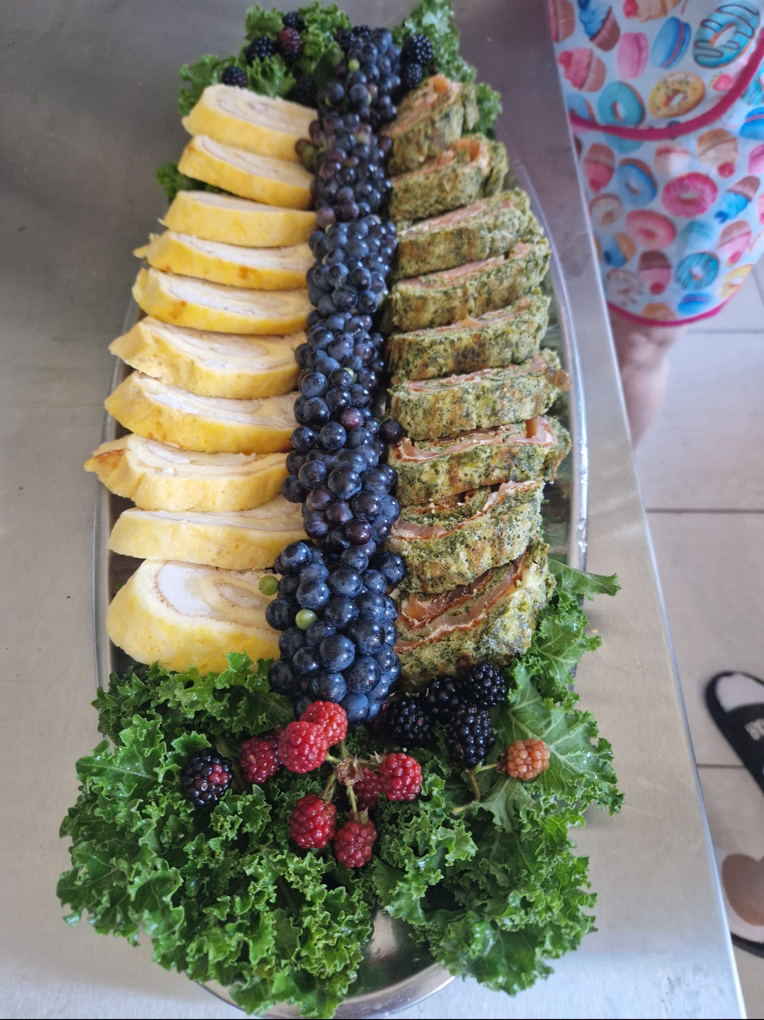 Dekoracyjna taca z roladkami jajecznymi, roladkami z łososia i szpinaku, ciemnymi winogronami, jeżynami i malinami na podkładzie z zielonego jarmużu.