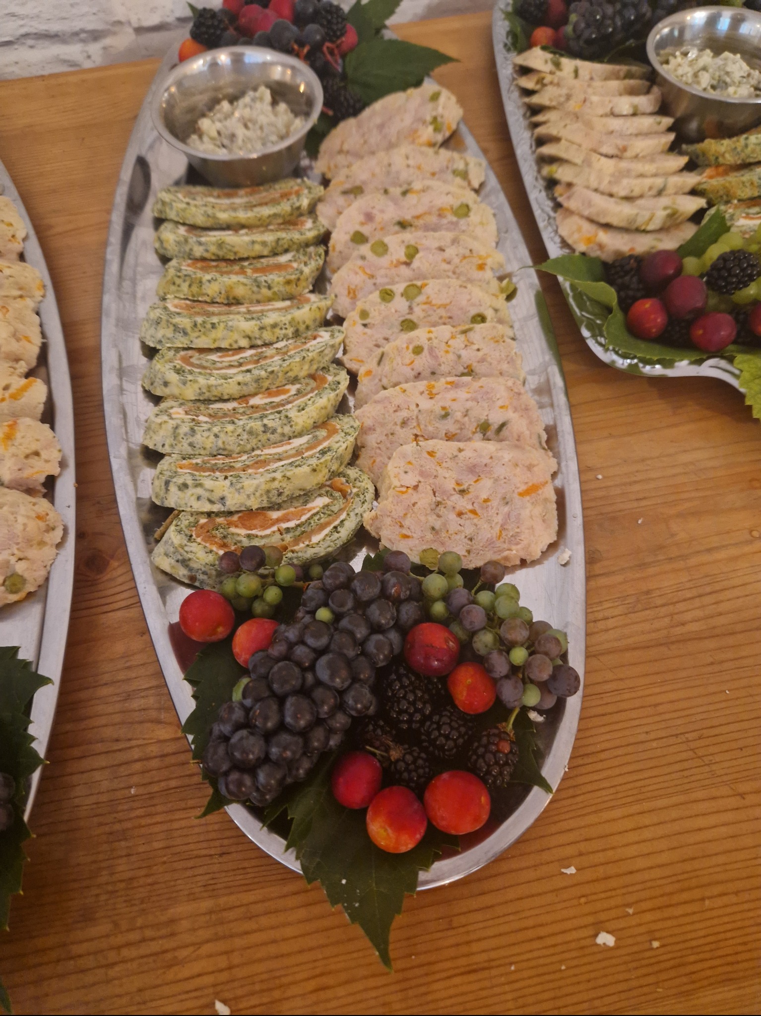 Dwa srebrne półmiski z przekąskami: roladki szpinakowe z łososiem, pasztet z warzywami, sos w metalowej miseczce oraz dekoracja z ciemnych i zielonych winogron, jeżyn i czerwonych śliwek...