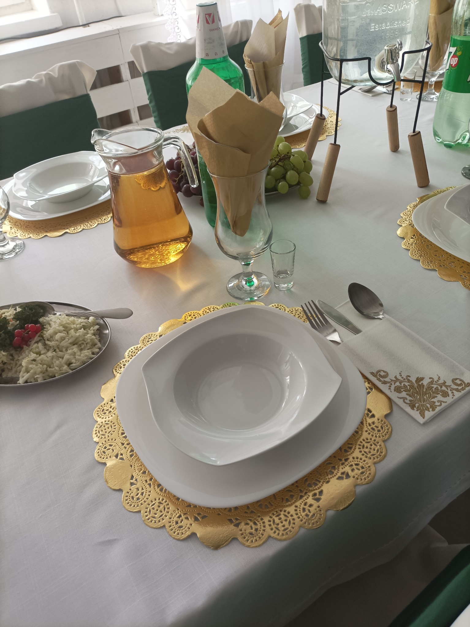 Elegancko nakryty stół z białym obrusem, złotymi podkładkami pod talerze, białymi naczyniami, dzbankiem z napojem, sałatką, winogronami i butelkami napojów.