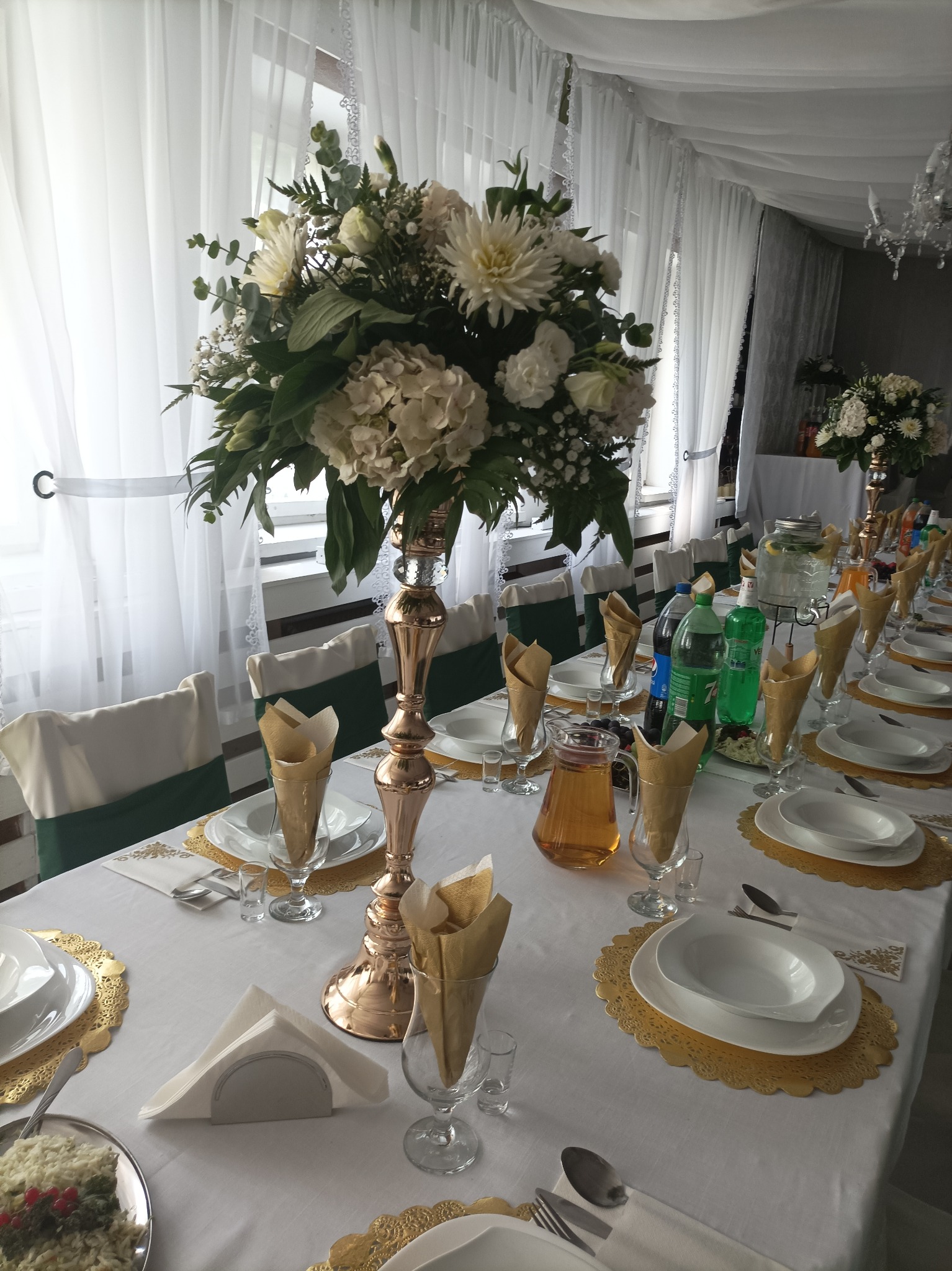 Uroczysty stół z białym obrusem, złotymi podkładkami, elegancką zastawą, kwiatowymi dekoracjami w wysokich, złotych wazonach oraz napojami. Krzesła z zielonymi akcentami. W tle białe, zwiewne firany.