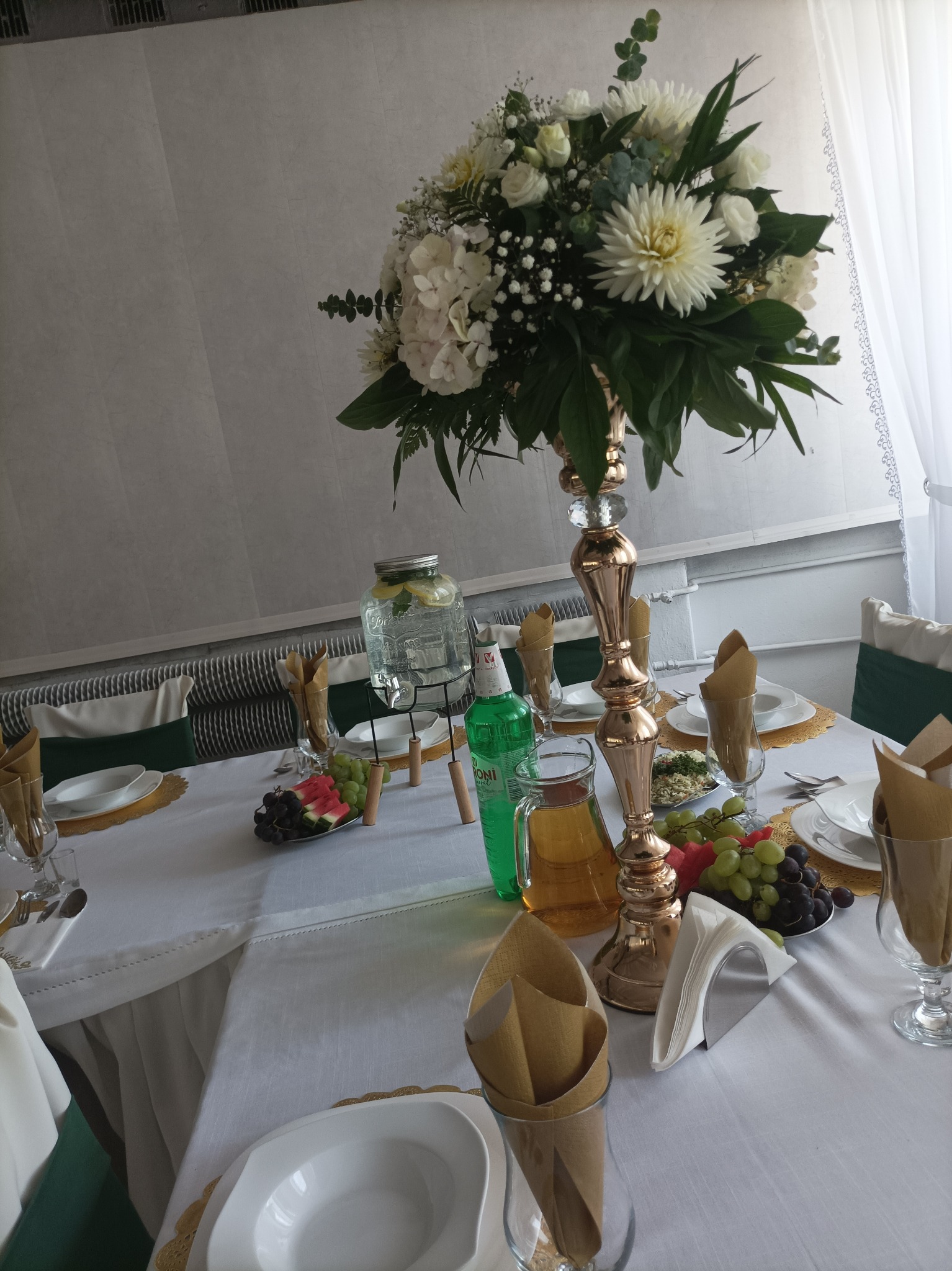 Elegancko nakryty stół z białym obrusem, złotymi podkładkami, wysokim wazonem z białymi kwiatami, szklanymi naczyniami z napojami, owocami i serwetkami w szklankach, przygotowany na przyjęcie.