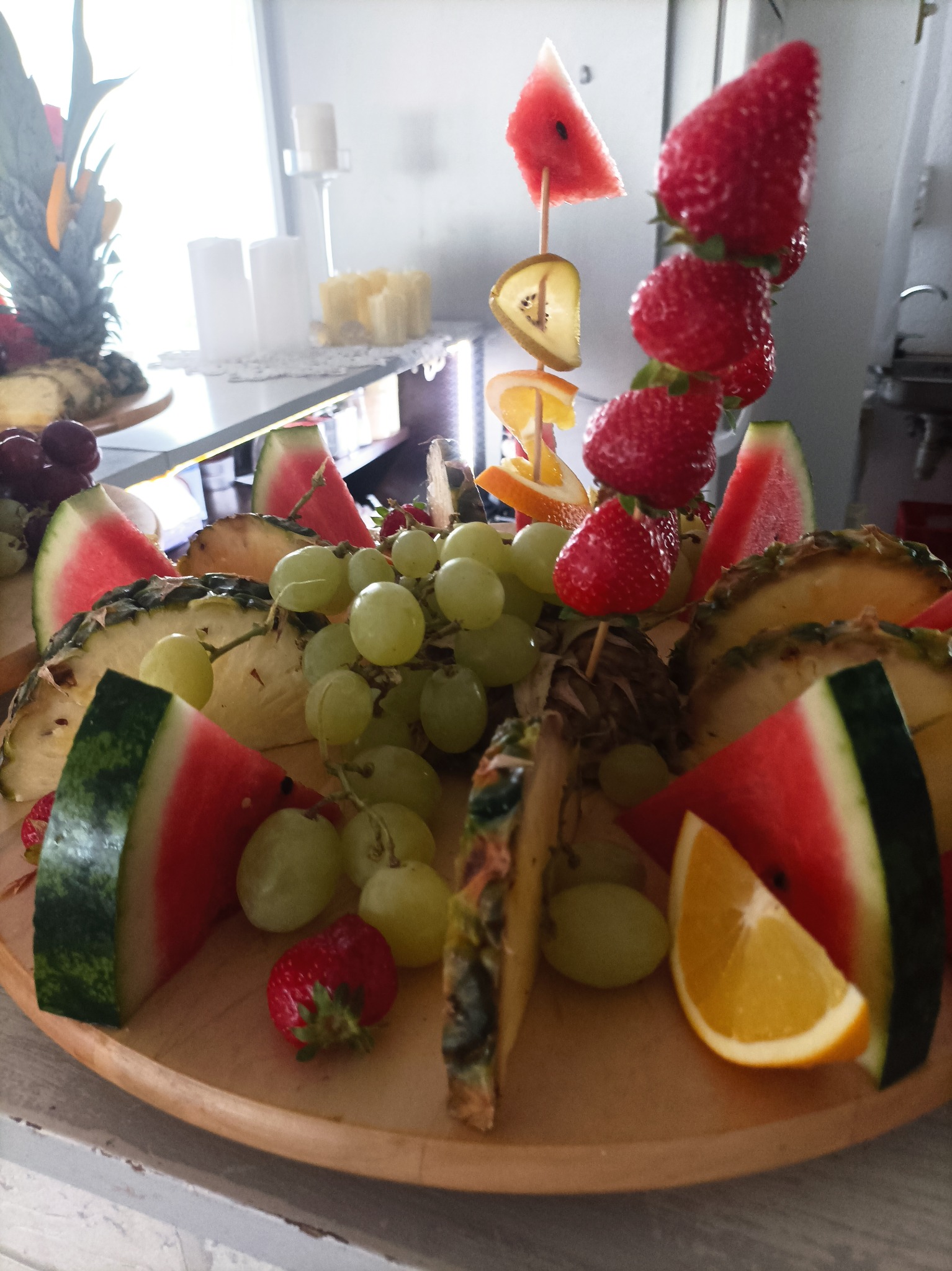 Dekoracyjny talerz z owocami: arbuz, ananas, winogrona, pomarańcze i szaszłyki z truskawek, kiwi i pomarańczy, na tle świec i dekoracji.