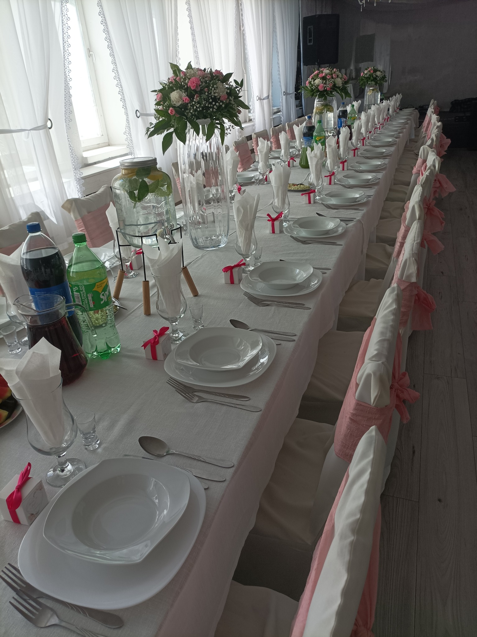 Długi stół bankietowy, nakryty białym obrusem, z białymi talerzami, sztućcami, szklankami, dzbankami z napojami i dekoracjami kwiatowymi w wysokich wazonach. Krzesła udekorowane różową wstążką.