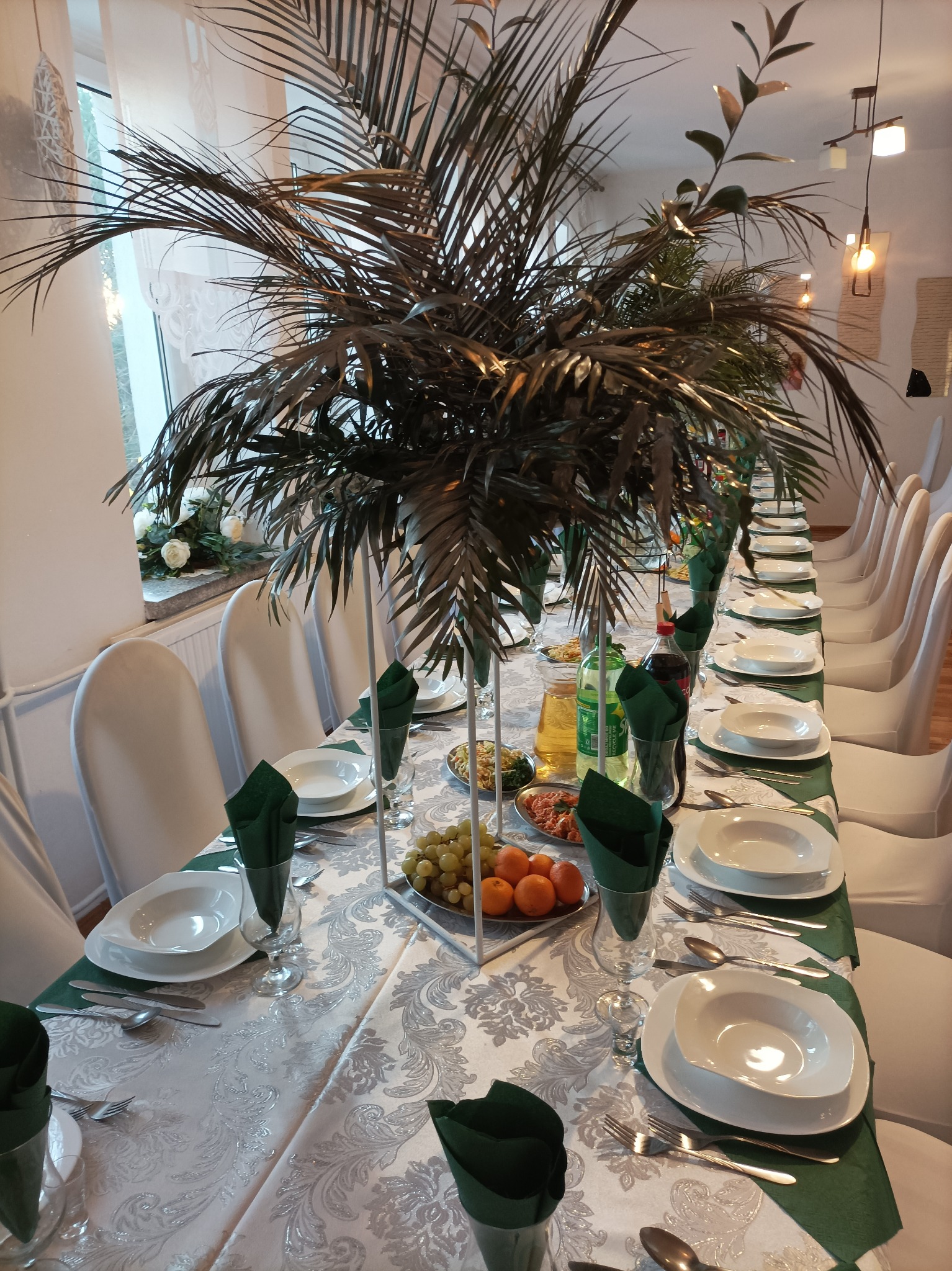 Uroczysty stół z białym obrusem w srebrne wzory, nakryty białymi talerzami, sztućcami, zielonymi serwetkami w szklankach, owocami i napojami, udekorowany wysoką kompozycją z gałęzi przypominających...