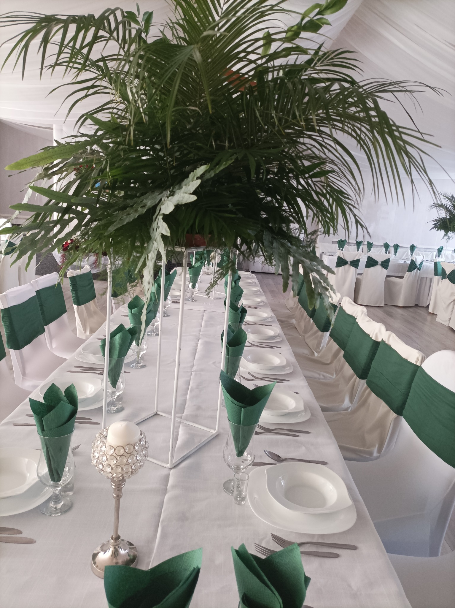 Uroczysty stół z białym obrusem, nakryciami i zielonymi serwetkami, ozdobiony wysokimi kompozycjami z tropikalnych liści i srebrnymi świecznikami, krzesła z zielonymi szarfami.