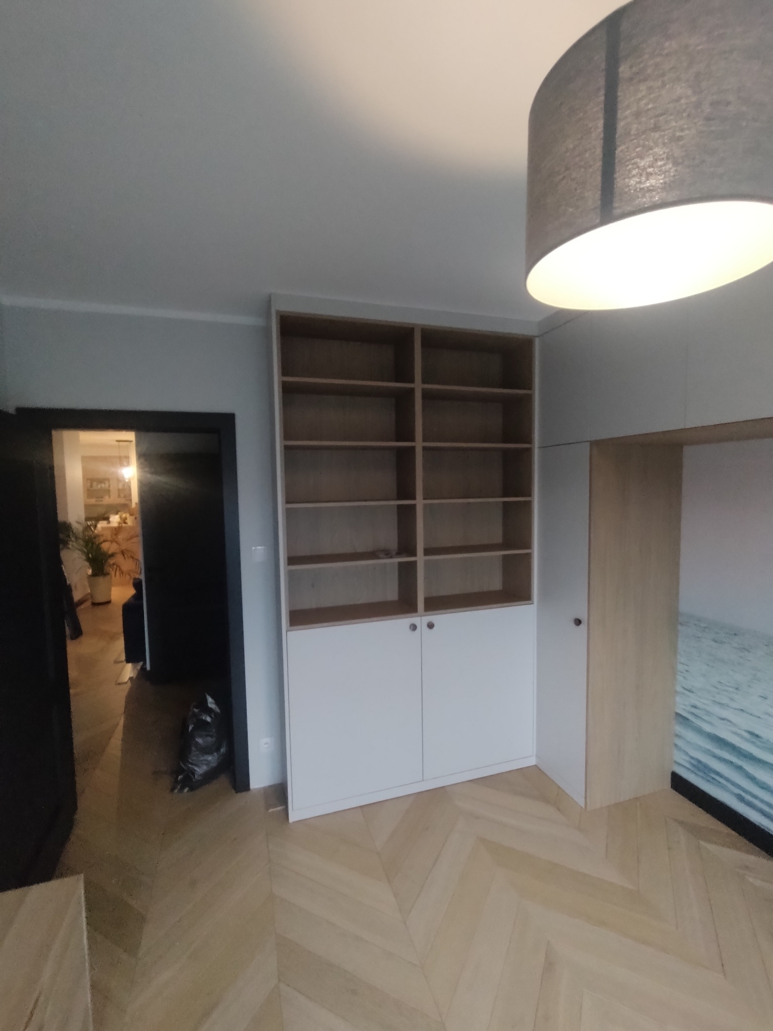 Wysoka, biała szafa z drewnianymi półkami, wbudowana w ścianę, z widocznymi drzwiami do innych pomieszczeń i parkietem ułożonym w jodełkę.