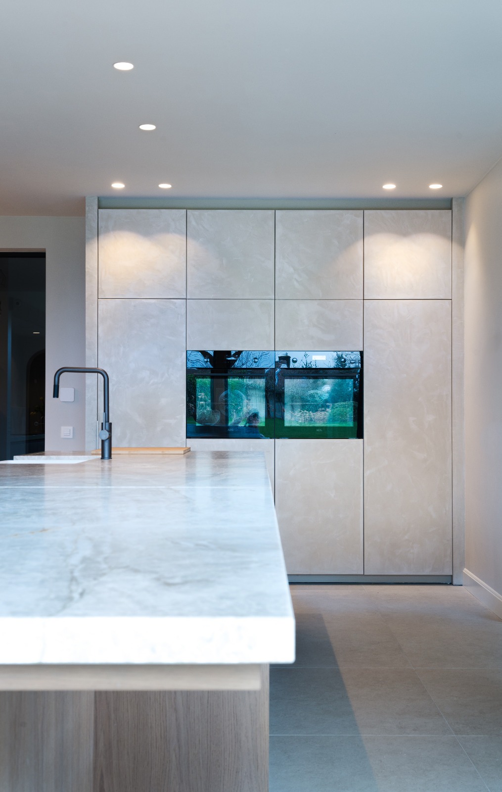 Nowoczesna kuchnia: zabudowa z marmurowymi frontami, wbudowane piekarniki, czarna bateria przy marmurowym blacie wyspy.