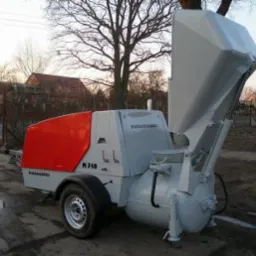 Mobilna pompa do betonu Putzmeister M740 z otwartym koszem zasypowym, zaparkowana na uboczu drogi, w tle widoczne domy jednorodzinne i drzewa bez liści.