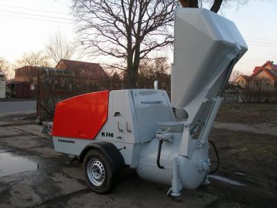 Mobilna pompa do betonu Putzmeister M740 z otwartym koszem zasypowym, zaparkowana na uboczu drogi, w tle widoczne domy jednorodzinne i drzewa bez liści.
