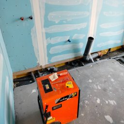 Nowacki - Niedokończona łazienka z niebieskimi płytami gipsowo-kartonowymi, częściowo pokrytymi gładzią, z widocznymi rurami i pomarańczowym osuszaczem Lehmann 50L z narzędziami na górze.