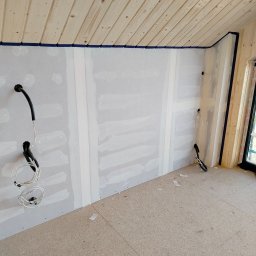 Nowacki - Wnętrze w trakcie prac wykończeniowych: ściany z płyt gipsowo-kartonowych z widocznymi łączeniami, zabezpieczone taśmą malarską, drewniana boazeria na skośnym suficie, wystające przewody elektryczne.