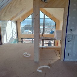 Nowacki - Wnętrze domu w trakcie wykańczania, widoczne drewniane panele na suficie, duże okna z widokiem na jezioro, ściany przygotowane do gładzi, podłoga z płyt OSB i resztki taśmy malarskiej.