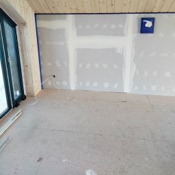 Nowacki - Wnętrze w trakcie prac wykończeniowych: zagruntowane płyty gipsowo-kartonowe na ścianach z widocznymi łączeniami wypełnionymi gładzią, zabezpieczone niebieską taśmą malarską, drewniana boazeria...