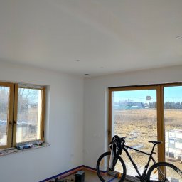 Nowacki - Wnętrze pokoju w trakcie remontu, z zabezpieczoną podłogą taśmą malarską, świeżo pomalowane ściany, rower oparty o okno oraz widok na zewnątrz.