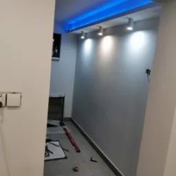Wnętrze w trakcie prac wykończeniowych z podświetlonym sufitem, widoczne płytki podłogowe, poziomica i lampy punktowe.
