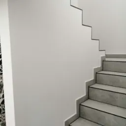 Minimalistyczne schody z szarych płytek z czarnymi akcentami, przylegające do białej ściany z dekoracyjnym wykończeniem, kontrastujące z czarno-białą tapetą w tle.