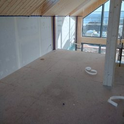 Nowacki - Wnętrze poddasza w trakcie wykańczania: surowe drewniane belki stropowe, ściany przygotowane do gładziowania z niebieską taśmą malarską, podłoga z płyt OSB, duże okno z widokiem na krajobraz.